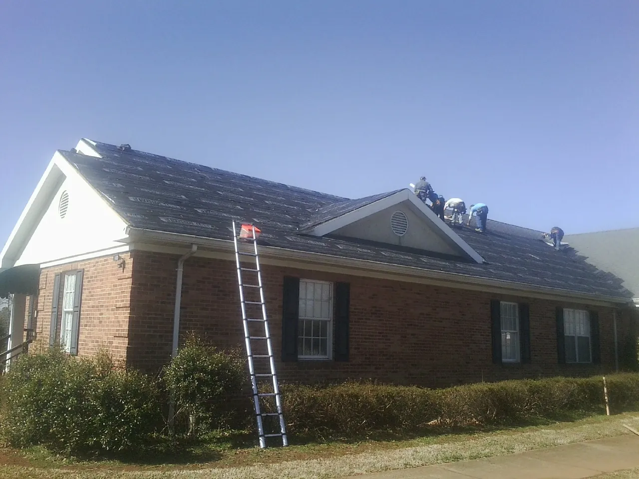 Crew tearing shingles off a brick ranch roof with a ladder set — Metal Roof Repair work by our crew in Duck Creek Village