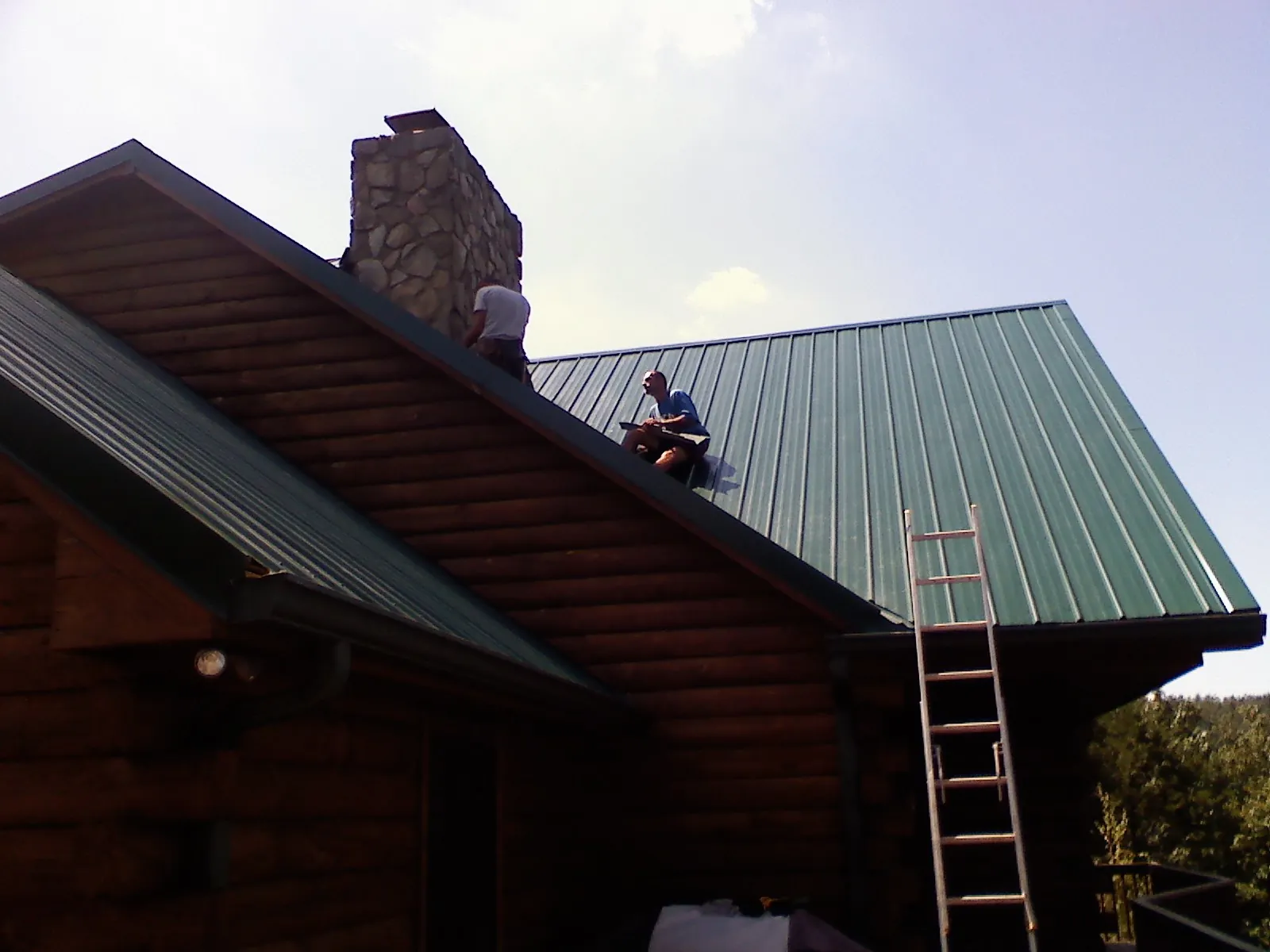 Roofers installing green metal panels beside a stone chimney on a log home in Duck Creek Village